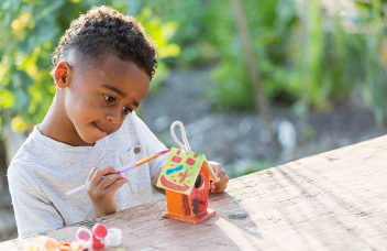 Here's an alt tag for the image: Young boy painting a birdhouse outdoors.