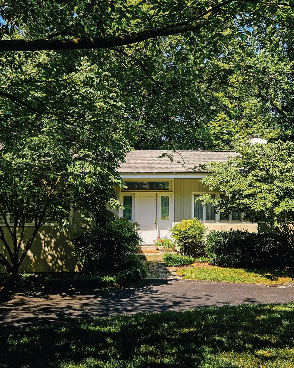 Small beige house with white door, surrounded by lush green trees